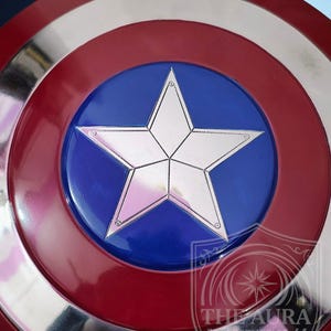 May include: A circular shield in red, white, and blue. The design includes a silver star on a blue central disc, surrounded by a silver band and a red outer ring. The shield's design is reminiscent of a superhero's emblem.