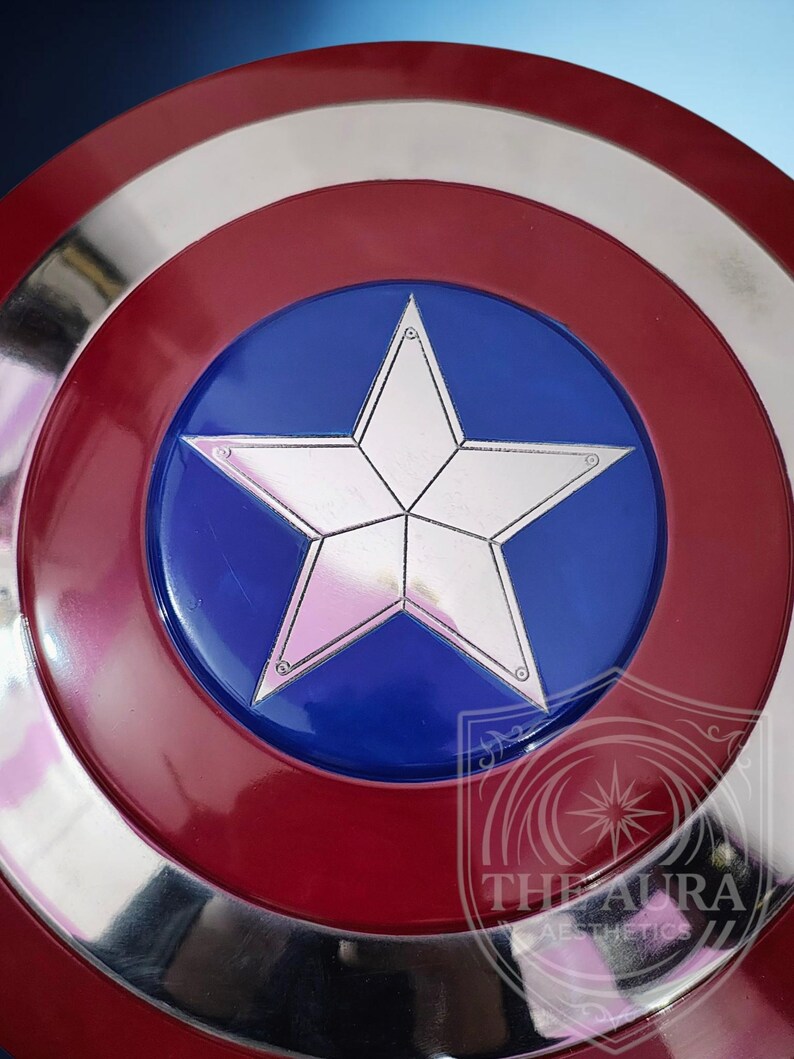 May include: A circular shield in red, white, and blue. The design includes a silver star on a blue central disc, surrounded by a silver band and a red outer ring. The shield's design is reminiscent of a superhero's emblem.