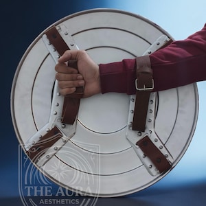 May include: A silver-coloured, round shield with brown leather straps and rivets. The shield is held by a person wearing a maroon long-sleeve shirt. The shield has a white centre and silver outer ring.