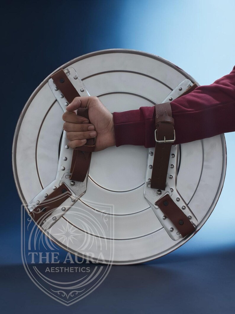 May include: A silver-coloured, round shield with brown leather straps and rivets. The shield is held by a person wearing a maroon long-sleeve shirt. The shield has a white centre and silver outer ring.