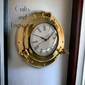 Könnte beinhalten: Eine goldfarbene Messinguhr mit römischen Ziffern auf einem weißen Zifferblatt. Die Uhr hat die Form eines runden Bullauges mit einem Messingrahmen und einem Messinggriff. Die Uhr ist an einer weißen Wand montiert.