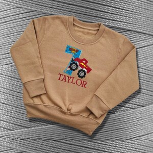 K&ouml;nnte beinhalten: Ein braunes Sweatshirt mit einem roten Monstertruck und der Nummer 7, die auf der Vorderseite gestickt sind. Der Text "TAYLOR" ist ebenfalls auf der Vorderseite des Sweatshirts gestickt.