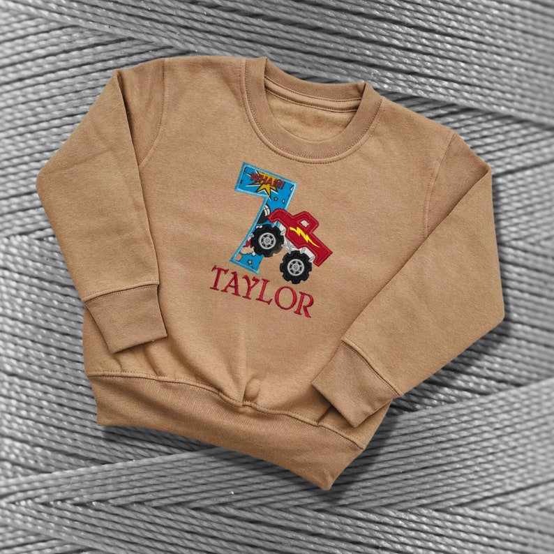 K&ouml;nnte beinhalten: Ein braunes Sweatshirt mit einem roten Monstertruck und der Nummer 7, die auf der Vorderseite gestickt sind. Der Text "TAYLOR" ist ebenfalls auf der Vorderseite des Sweatshirts gestickt.