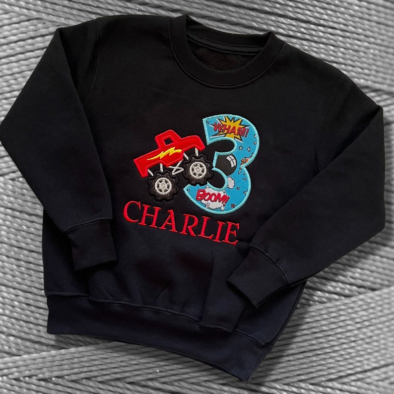 K&ouml;nnte beinhalten: Schwarzer Pullover mit einem roten Monstertruck und der Zahl 3, die auf der Vorderseite gestickt ist. Die Zahl 3 ist von blauen und wei&szlig;en Wolken mit den Worten "WHAM!" und "BOOM!" umgeben. Der Name "CHARLIE" ist unter dem Truck gestickt.