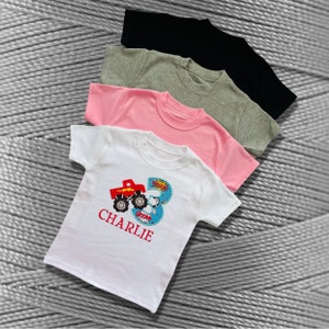 K&ouml;nnte beinhalten: Ein wei&szlig;es T-Shirt mit einem roten Monstertruck und der Zahl 3. Das Shirt hat auch das Wort "BOOM" und den Namen "CHARLIE" aufgedruckt.