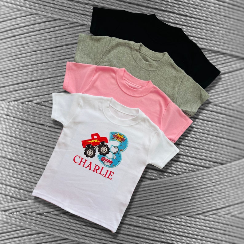 K&ouml;nnte beinhalten: Ein wei&szlig;es T-Shirt mit einem roten Monstertruck und der Zahl 3. Das Shirt hat auch das Wort "BOOM" und den Namen "CHARLIE" aufgedruckt.