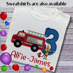 May include: A white t-shirt featuring an embroidered red fire engine, a large blue glittery number three, and a firefighter figure holding a hose. The name Alfie-James is embroidered in red below the graphic. Text at the top reads: Sweatshirts are also available.