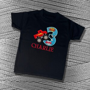 K&ouml;nnte beinhalten: Schwarzes T-Shirt mit einem roten Monstertruck und der Zahl 3 in Blau mit wei&szlig;em Umriss. Die Zahl 3 hat einen gelben Stern und die Worte "WHAM!" und "BOOM!" darauf. Der Name "CHARLIE" ist in Rot unter der Zahl gestickt.