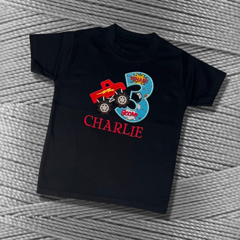 K&ouml;nnte beinhalten: Schwarzes T-Shirt mit einem roten Monstertruck und der Zahl 3 in Blau mit wei&szlig;em Umriss. Die Zahl 3 hat einen gelben Stern und die Worte "WHAM!" und "BOOM!" darauf. Der Name "CHARLIE" ist in Rot unter der Zahl gestickt.