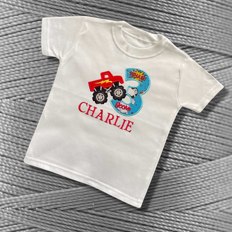 K&ouml;nnte beinhalten: Wei&szlig;es T-Shirt mit einem roten Monstertruck und einer blauen Zahl 3 mit den Worten "BOOM!" und "WHAAM!" darauf. Der Name "CHARLIE" ist in Rot unter dem Truck gestickt.
