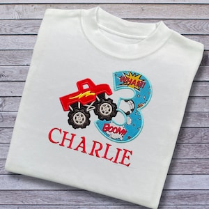 K&ouml;nnte beinhalten: Wei&szlig;es T-Shirt mit einem roten Monstertruck und einer blauen Zahl 3 mit den Worten "WHAM!" und "BOOM!" darauf. Der Name "CHARLIE" ist in Rot unter dem Design gestickt.