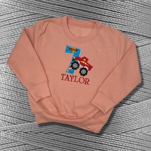 K&ouml;nnte beinhalten: Ein pfirsichfarbener Pullover mit einem roten Monstertruck-Design und der Zahl 7. Der Truck hat eine blaue und wei&szlig;e "POW"-Grafik. Der Name "TAYLOR" ist unter dem Truck gestickt.