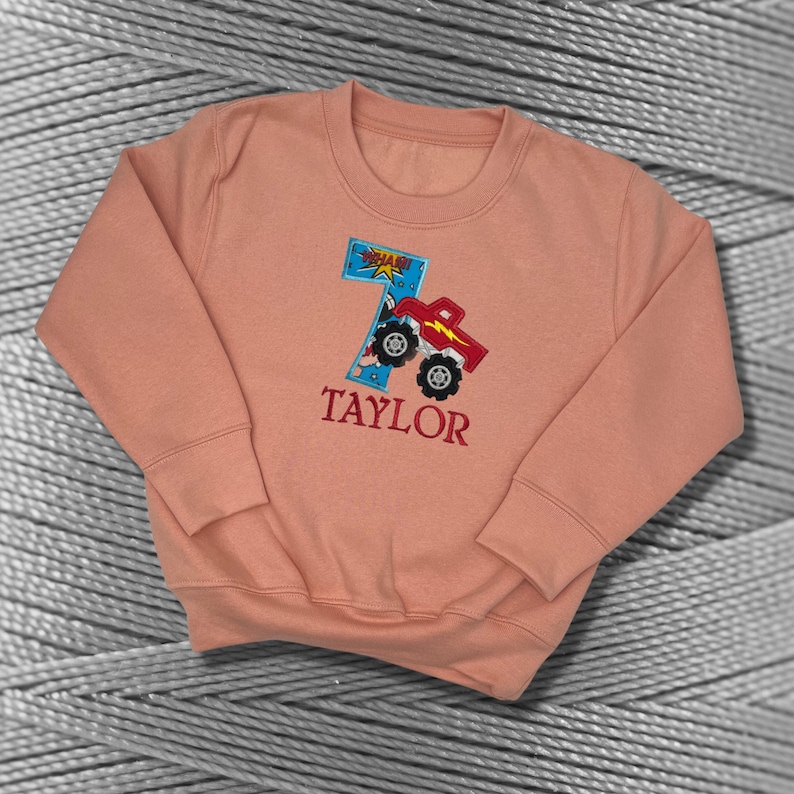 K&ouml;nnte beinhalten: Ein pfirsichfarbener Pullover mit einem roten Monstertruck-Design und der Zahl 7. Der Truck hat eine blaue und wei&szlig;e "POW"-Grafik. Der Name "TAYLOR" ist unter dem Truck gestickt.