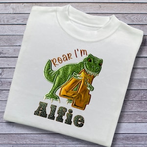 May include: White t-shirt with a green dinosaur holding a gold number 4. The text "Roar I'm Alfie" is embroidered on the shirt.