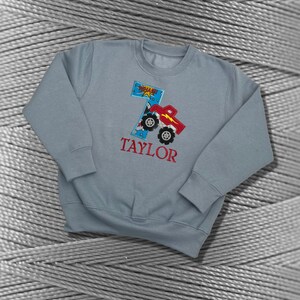 K&ouml;nnte beinhalten: Ein hellblauer Pullover mit einem roten Monstertruck-Design und der Zahl 7. Der Text "TAYLOR" ist unter dem Truck gestickt.