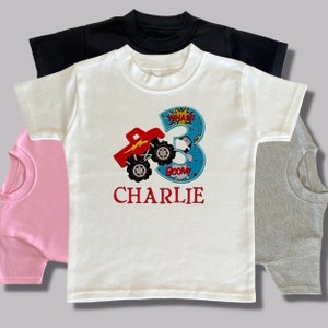 K&ouml;nnte beinhalten: Wei&szlig;es T-Shirt mit einem roten Monstertruck und einem blauen Nummer 3 Applique. Das Applique hat die Worte "WHAM!" und "BOOM!" drauf. Der Name "CHARLIE" ist in Rot unter dem Applique gestickt.