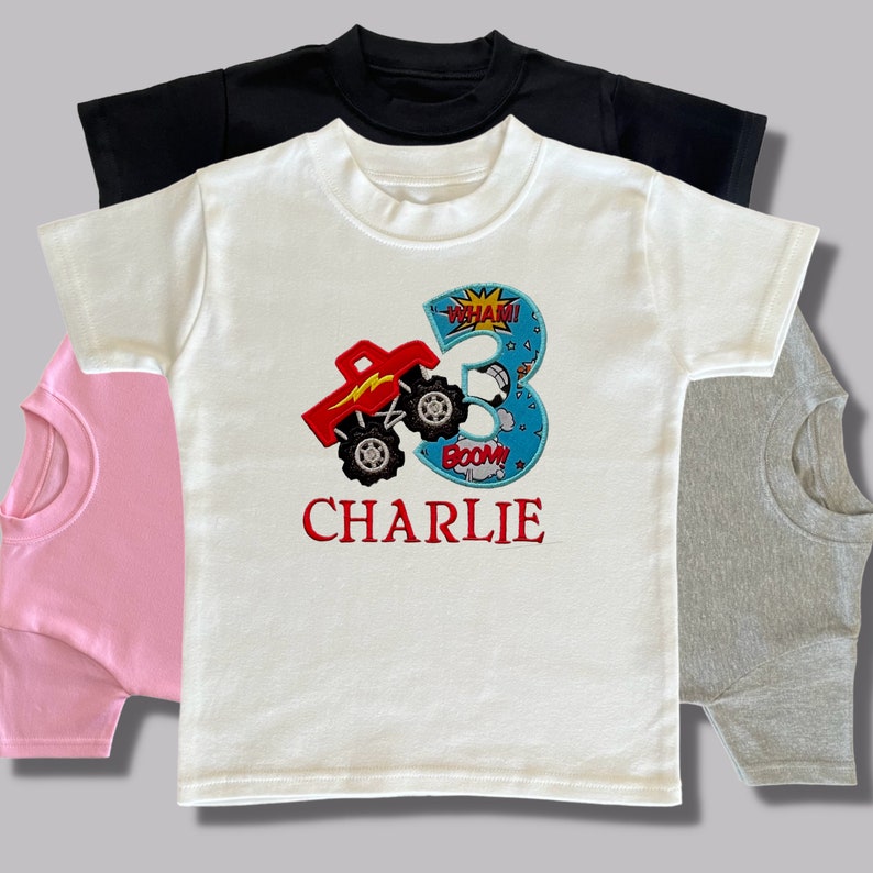K&ouml;nnte beinhalten: Wei&szlig;es T-Shirt mit einem roten Monstertruck und einem blauen Nummer 3 Applique. Das Applique hat die Worte "WHAM!" und "BOOM!" drauf. Der Name "CHARLIE" ist in Rot unter dem Applique gestickt.