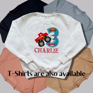 K&ouml;nnte beinhalten: Wei&szlig;er Pullover mit einem roten Monstertruck und der Nummer 3 darauf. Der Text "WHAM!" und "BOOM!" befindet sich auf dem Truck. Der Name "CHARLIE" ist unter dem Truck gestickt. T-Shirts sind ebenfalls erh&auml;ltlich.