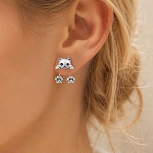 May include: A silver-toned stud earring featuring a cat head design with black eyes and nose, with two dangling silver-toned paw print charms suspended from the cat's front paws, creating a layered look on the earlobe.