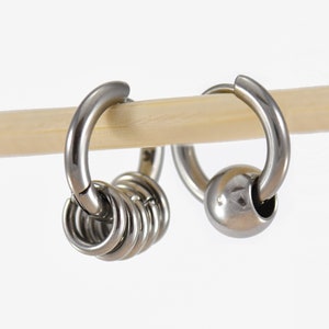 May include: Two silver hoop earrings. One has a small ball, the other a series of small rings. The earrings are displayed on a light-coloured wooden dowel against a white background.