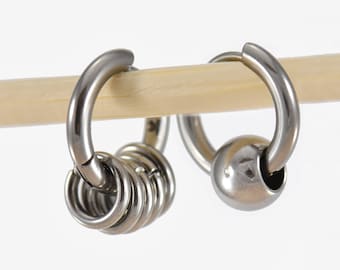 May include: Two silver-colored hoop earrings. One earring features a small ball, while the other has a series of small rings. The earrings are displayed on a light-colored wooden dowel against a white background.