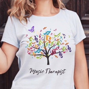 Könnte beinhalten: Ein hellblaues T-Shirt mit einem Motiv eines Baumes aus Musiknoten. Ein lilafarbener Schmetterling fliegt über dem Baum. Der Text "Music Therapist" ist unter dem Baum gedruckt.