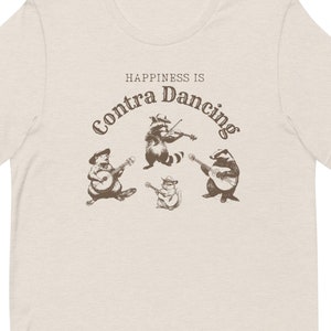 May include: A beige t-shirt featuring a brown graphic of raccoons playing musical instruments. The text "Happiness is Contra Dancing" is printed on the shirt.