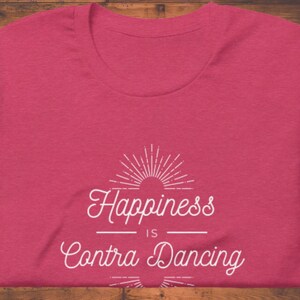 May include: Pink t-shirt with a white graphic that says "Happiness is Contra Dancing" with a sunburst design above the text.