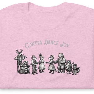 May include: A light pink t-shirt featuring a black and white ink-style illustration of anthropomorphic forest animals dancing and playing music. The text "CONTRA DANCE JOY" is printed in a curved arch above the group of animals in a grey font.