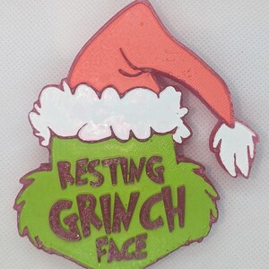 May include: A Christmas-themed decoration featuring a green Grinch face with the words "RESTING GRINCH FACE" in a darker green font. The Grinch face is topped with a red Santa hat with white trim.