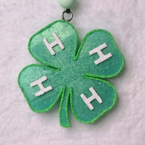 May include: A green four-leaf clover pendant with white glitter trim and white "H" letters on each leaf. The pendant is attached to a black cord with a light green bead and silver ring.