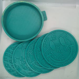 May include: A teal coaster set with a matching holder. The coasters feature festive designs, including snowflakes, candy canes, and smiling faces with Christmas stockings. The holder has a handle for easy carrying.