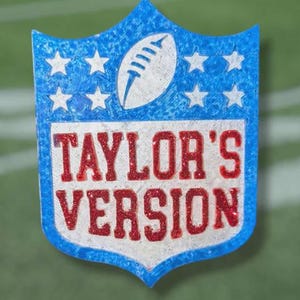 May include: A blue, white, and red shield-shaped emblem with the text "TAYLOR'S VERSION." The emblem features a football and stars, resembling a sports league logo. The background is a green field.