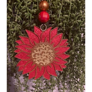 May include: A red glitter sunflower ornament with a gold glitter leopard print center. The ornament is hanging from a string with two red and gold beads.