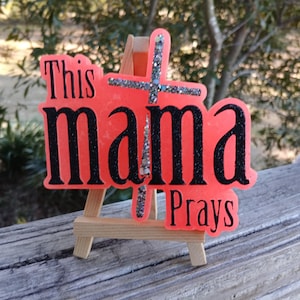 May include: A pink sign with the words "This mama prays" and a glittery silver cross in the center. The sign is on a small wooden easel.