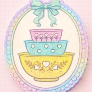 May include: An oval-shaped sticker featuring three pastel-colored mixing bowls stacked on top of each other. The top bowl is pink, the middle is blue, and the bottom is yellow. Each bowl has a different heart and floral design. A light blue bow sits above the bowls.