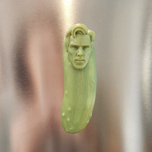 May include: A green pickle with a man's face carved into it. The face has a serious expression.