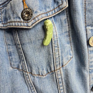 May include: A green pickle pin with a human face on a denim jacket.