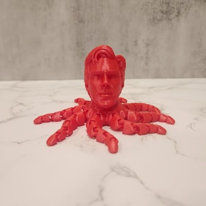 May include: A red 3D printed sculpture of a human head with a serious expression, attached to the body of an octopus.