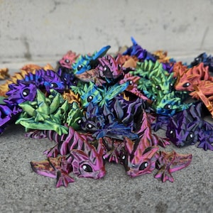 May include: A collection of colorful, 3D-printed dragon figurines in various shades of purple, pink, blue, green, and orange. Each dragon has a detailed, textured design with small, round eyes. The dragons are scattered on a gray surface.
