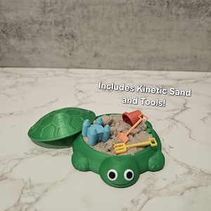 May include: A green turtle-shaped sandbox with a lid, filled with kinetic sand and small tools. The set includes a red bucket, yellow rake, and blue castle. Text on the image reads "Includes Kinetic Sand and Tools!" and "*colors of tools may vary."