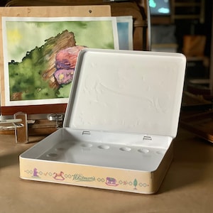 Könnte beinhalten: Eine offene weiße Metall-Farbpalette mit einem verspielten Design mit einem Schaukelpferd, einem Haus und einem Baum. Die Palette hat 8 runde Vertiefungen für Farbe und ist mit "Whitman's" beschriftet.