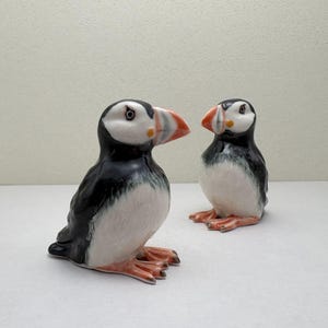 May include: Two ceramic puffin figurines. The birds have black backs, white bellies, and orange feet and beaks. The beaks have a touch of yellow and gray. One puffin is standing, and the other is sitting.