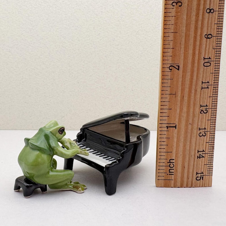 May include: A whimsical ceramic figurine of a green frog playing a black piano. The piano has white and black keys. The frog is seated on a small black stool. The image includes a ruler showing the piano is approximately 2 inches in height.