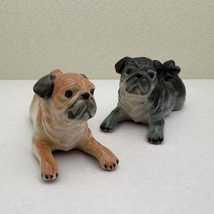 Könnte beinhalten: Zwei Keramik-Mops-Hunde-Figuren. Einer ist hellbraun mit schwarzen Akzenten, der andere ist grau mit schwarzen Akzenten. Beide liegen mit ausgestreckten Vorderpfoten.