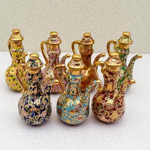 Könnte beinhalten: Eine Sammlung von Miniatur-Dekorations-Teekannen. Jede Teekanne hat einen bauchigen Körper, einen gebogenen Ausguss und einen Griff, die alle mit aufwendigen Blumenmustern in verschiedenen Farben verziert sind, darunter Gold, Blau, Rosa und Grün. Die Teekannen haben goldfarbene Akzente.