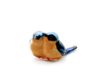 May include: A ceramic figurine of two bluebirds, facing each other, with yellow and orange markings. The birds are joined together at the chest, with one bird's tail extending to the side.