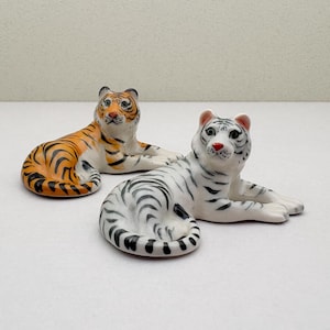 Op de afbeelding: Twee keramische tijgerbeeldjes. Eén is oranje en zwart, de andere is wit met zwarte strepen. Beide bevinden zich in een liggende houding, met gedetailleerde gelaatstrekken en realistische markeringen.