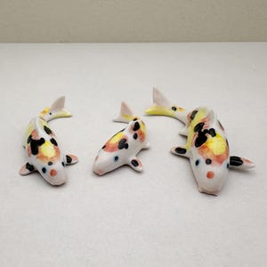 Pode incluir: Três estatuetas de peixe koi de cerâmica. Os peixes são pintados em tons de branco, laranja, amarelo e preto. Os peixes estão em várias poses, com suas caudas e barbatanas estendidas.