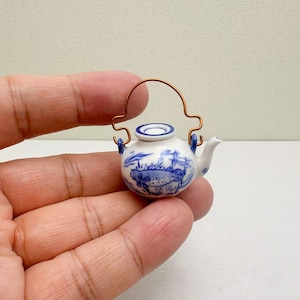 Può includere: Teiera in miniatura in porcellana bianca e blu con manico in filo di rame. La teiera presenta un disegno di pesce e fiori blu. Il beccuccio e gli attacchi del manico sono blu. La teiera è tenuta in mano.